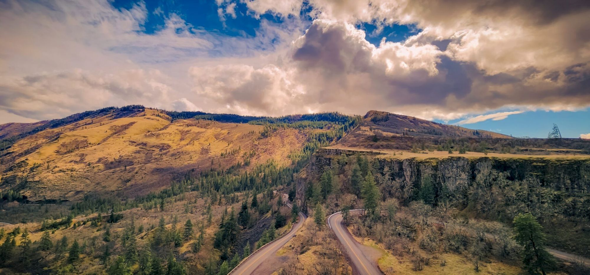 A winding road leads through a scenic mountain landscape with dramatic clouds and trees scattered across the hillsides.