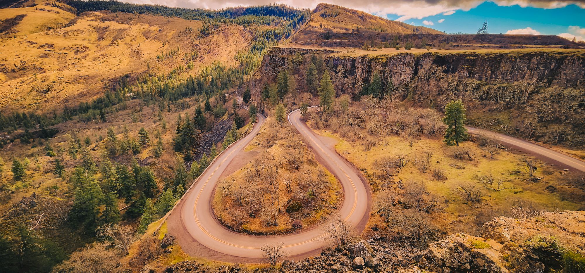 A winding road snakes through a hilly, forested landscape with steep cliffs and scattered trees under a partly cloudy sky.