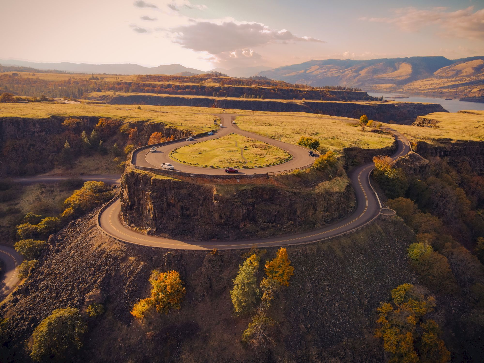 A curvy road loops around a plateau with scenic views, surrounded by autumn trees and hills under a cloudy sky.