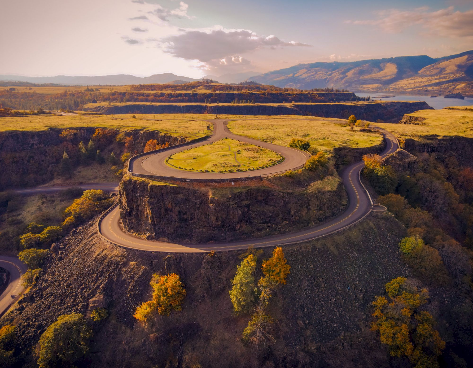Aerial view of a winding road on a plateau with scenic landscapes, surrounded by trees and hills under a cloudy sky at sunset.
