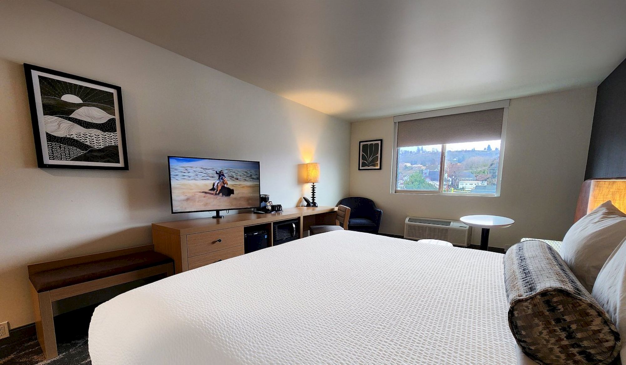 A hotel room features a bed, TV, artwork, lamp, armchair, and window with a view outside.