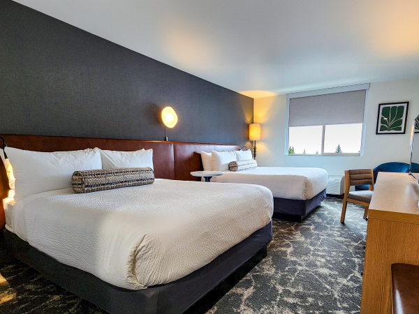 A hotel room with two beds, modern decor, wall art, and a TV on a wooden dresser by the window with natural light.