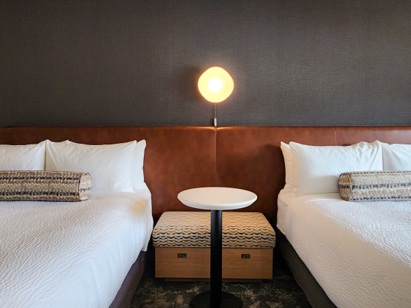 A hotel room with two neatly made beds, a central table, pillows, cushions, and bedside lamps on either side.