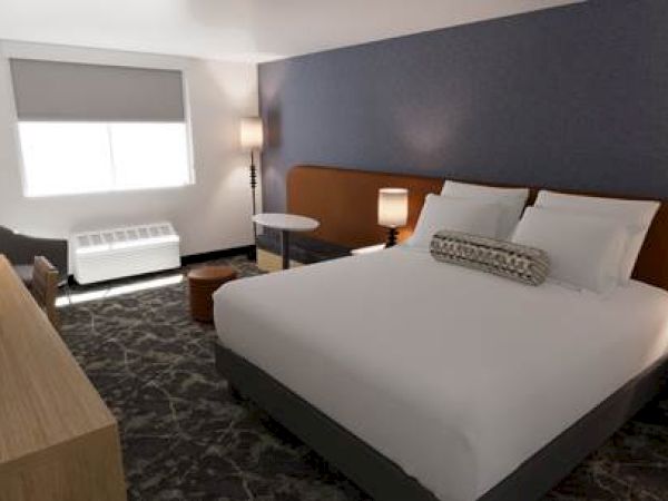 A hotel room features a large bed, bedside tables with lamps, a small table, an armchair, and a TV on a wooden dresser.