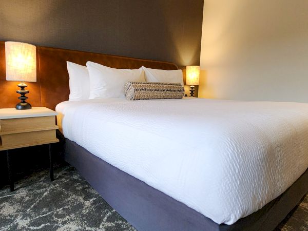 A neatly made bed with white linens sits between two bedside tables with lamps, in a modern, carpeted room.