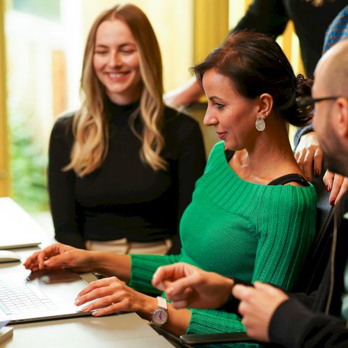 A group of people are gathered around a laptop, looking at the screen and smiling in a well-lit room.