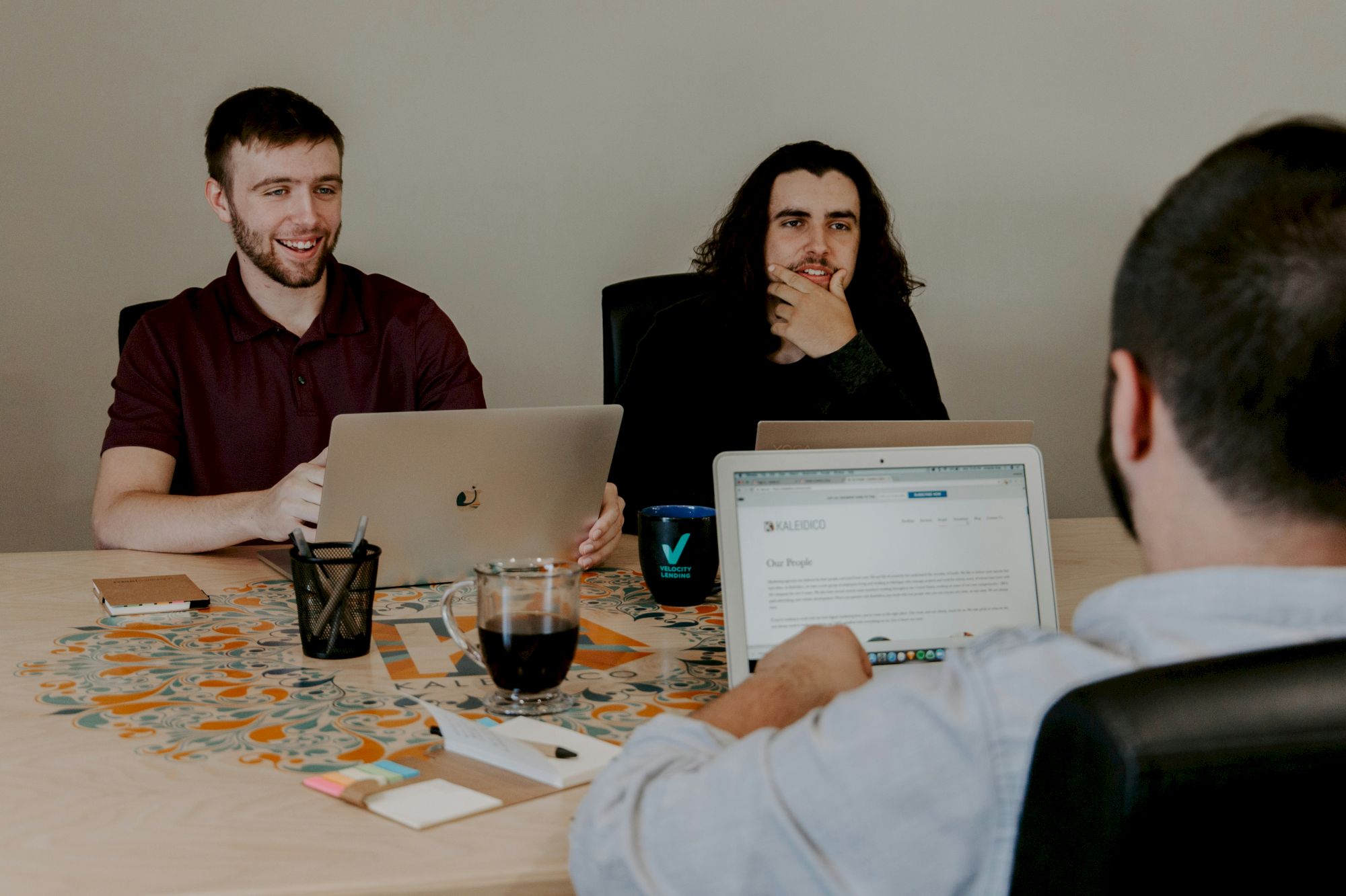 Three people are sitting around a table with laptops, engaged in discussion and appearing focused.