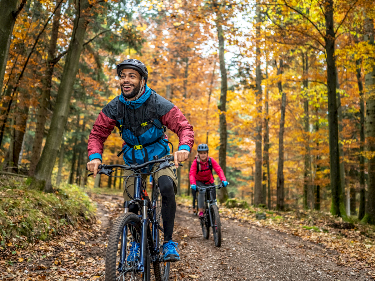 Two people are joyfully mountain biking on a forest trail surrounded by autumn foliage.