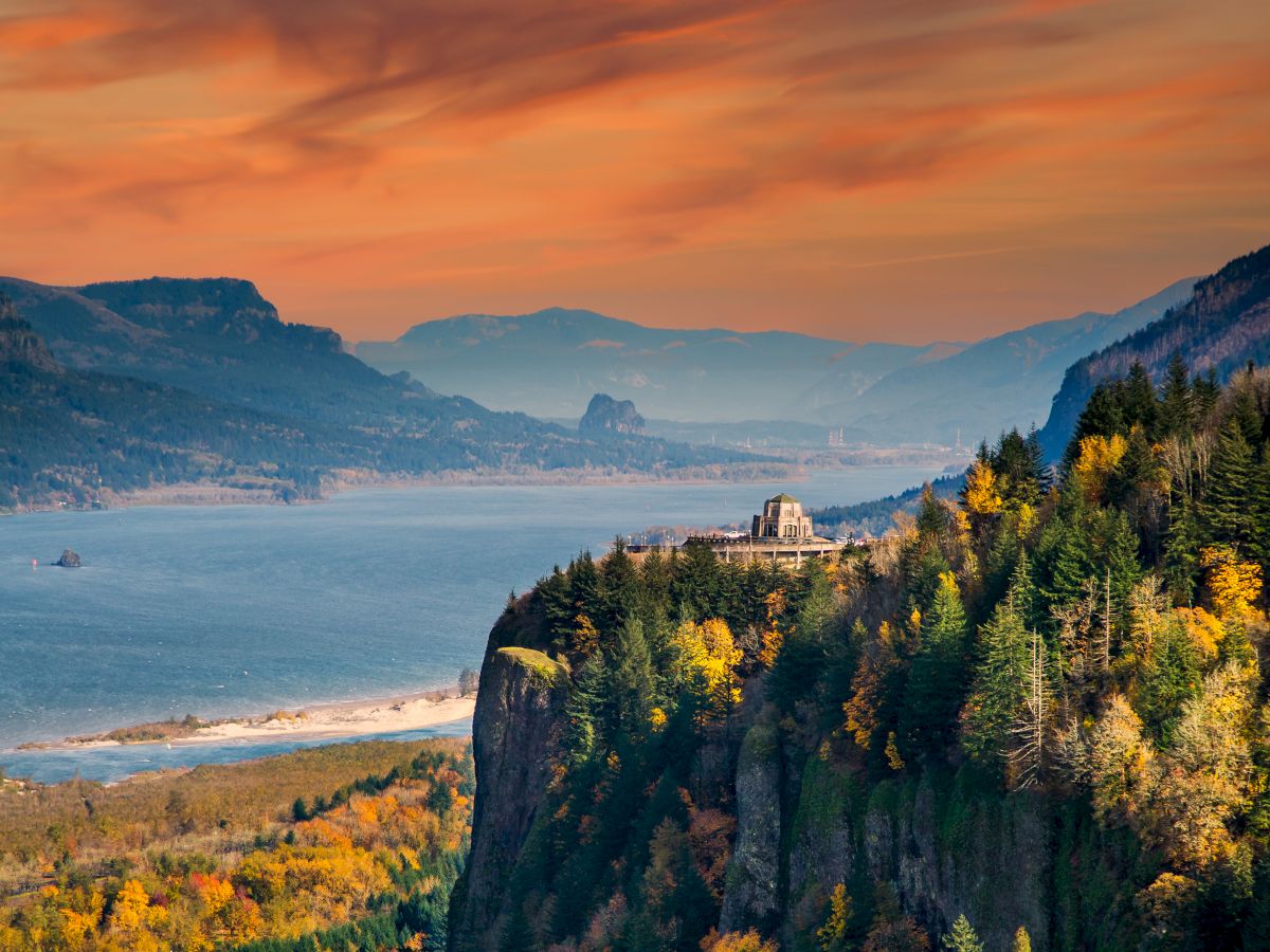 A scenic view of a river winding through a valley with autumn foliage, rocky cliffs, and a colorful sunset sky in the background.