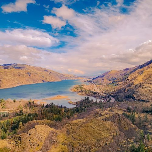 A vast desert valley with rocky hills, a winding road, and distant mountains under a dramatic blue sky with golden sunlight.