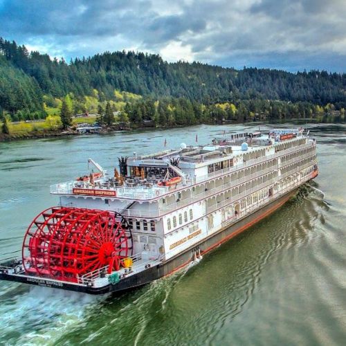 A red and white boat glides on blue-green water, with a green forest and hills in the background.