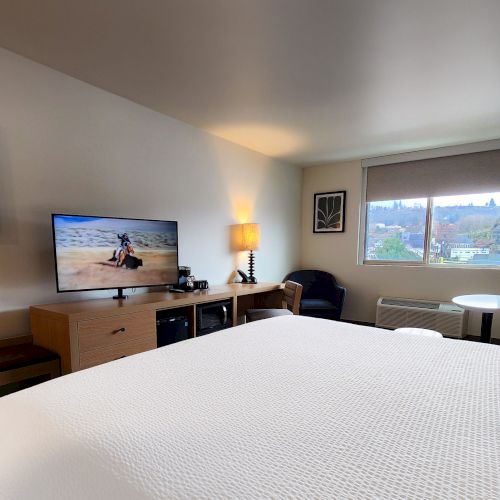 A cozy hotel bedroom with a large bed, wall-mounted TV, desk, seating area, and soft lighting in a modern, neutral-toned space.