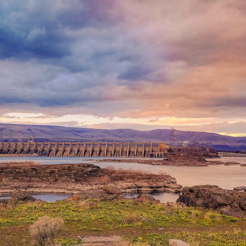 A scenic landscape with a river, a dam, rolling hills, and a dramatic sky during sunset or sunrise, giving a serene and vibrant atmosphere.