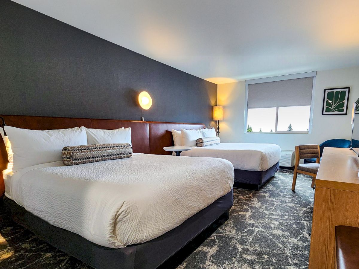 A modern hotel room with two neatly made double beds, a TV, a chair, a desk, and a window offering natural light.