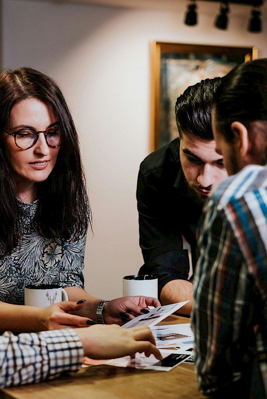 A group of four people sitting around a table, engaged in discussion and reviewing documents or photos, with coffee cups nearby.