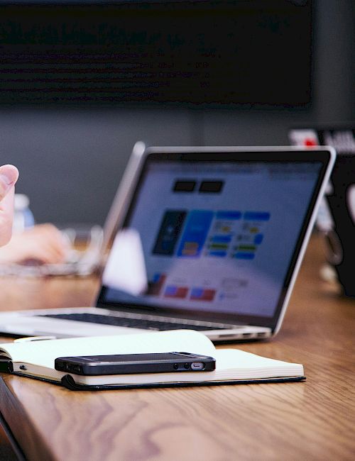A person gestures while seated at a table with a laptop, notebook, and smartphone, suggesting a meeting or discussion setting.