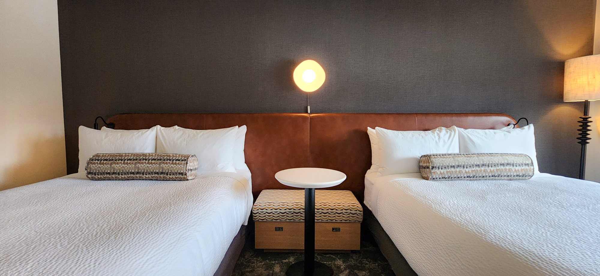 The image shows a hotel room with two neatly made beds, a round table, and a lamp, offering a cozy and well-organized atmosphere.